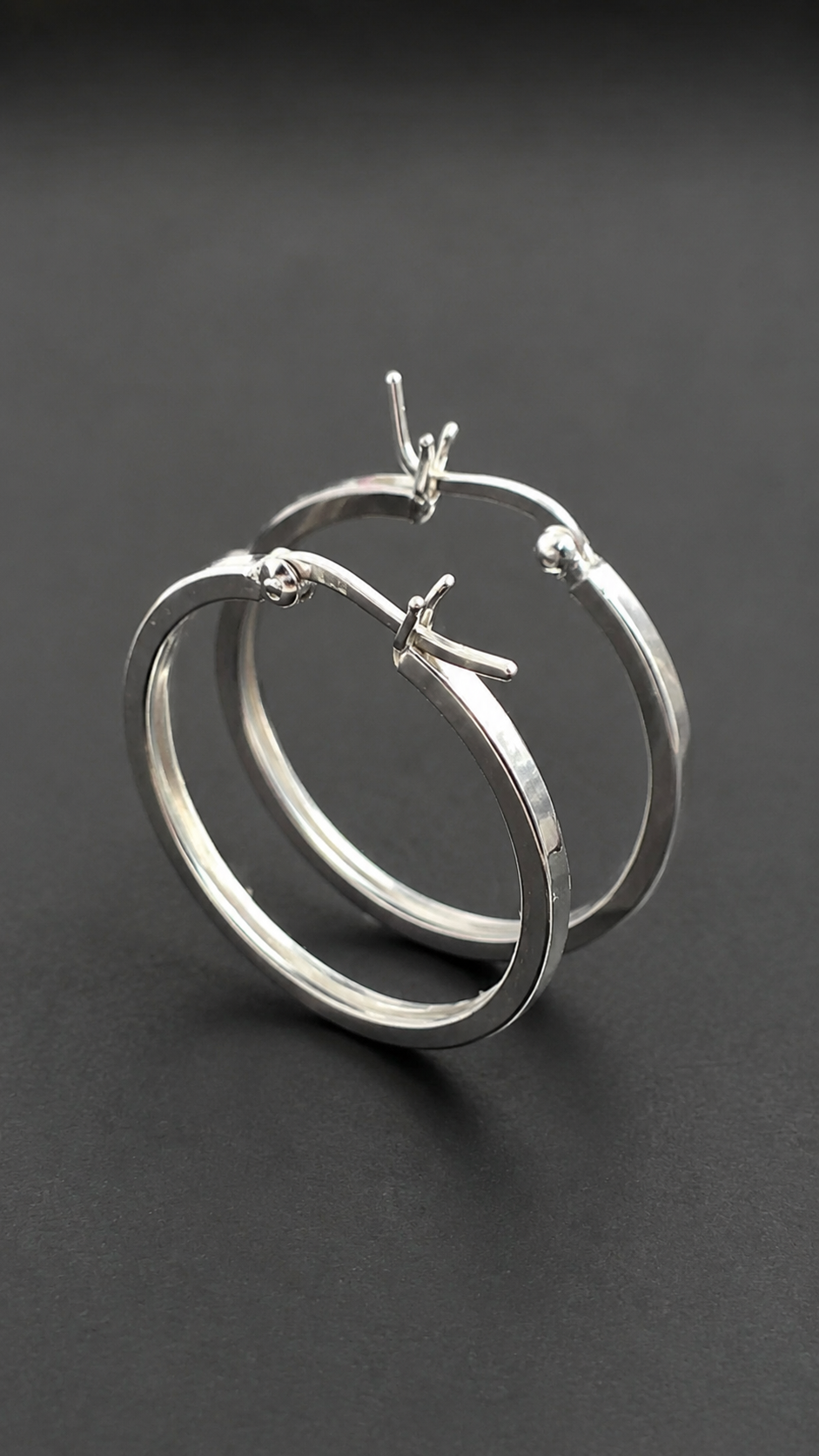 A pair of polished silver hoop earrings with small decorative wire details at the top and side, displayed on a dark gray background.
