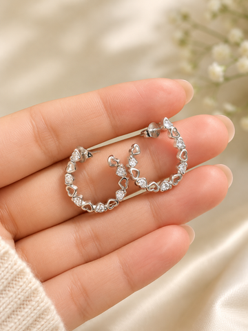 A pair of silver-tone hoop earrings with small clear heart-shaped stones, held in a person's hand against a soft beige satin and blurred floral background.