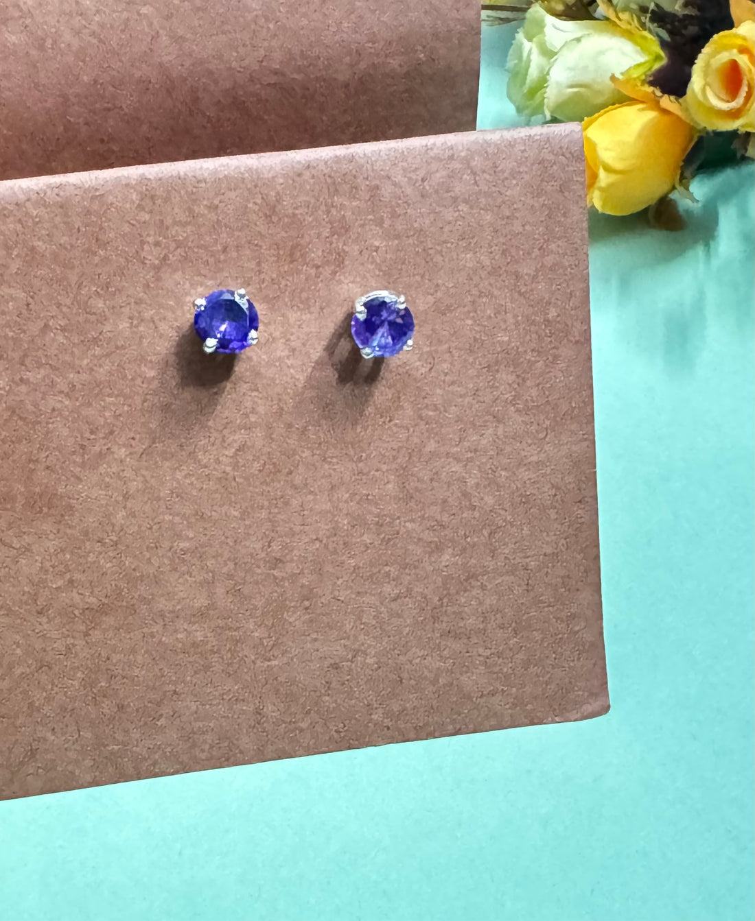 Pair of blue gemstone stud earrings on a brown card with flowers in the background.