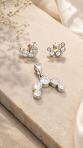Silver-toned jewelry set featuring a pendant and matching stud earrings with clear round gemstones, arranged on a beige stone slab with soft fabric in the background.