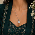 Person wearing a silver necklace with a 'B' pendant against a neutral background
