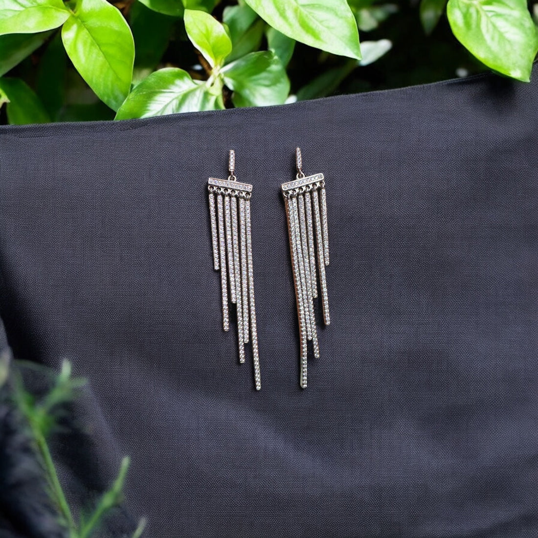 Silver fringe earrings on a black surface with green leaves in the background