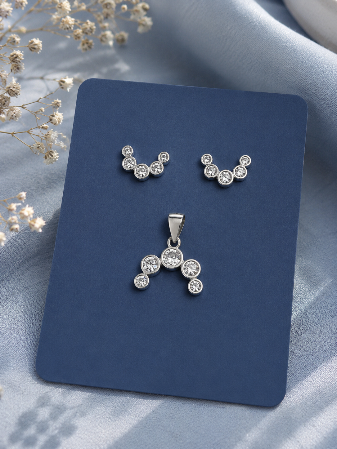 Silver-tone stud earrings and matching pendant set with clear round gemstones, displayed on a dark blue card against a soft light blue fabric background with dried flowers.