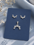 Silver-tone stud earrings and matching pendant set with clear round gemstones, displayed on a dark blue card against a soft light blue fabric background with dried flowers.