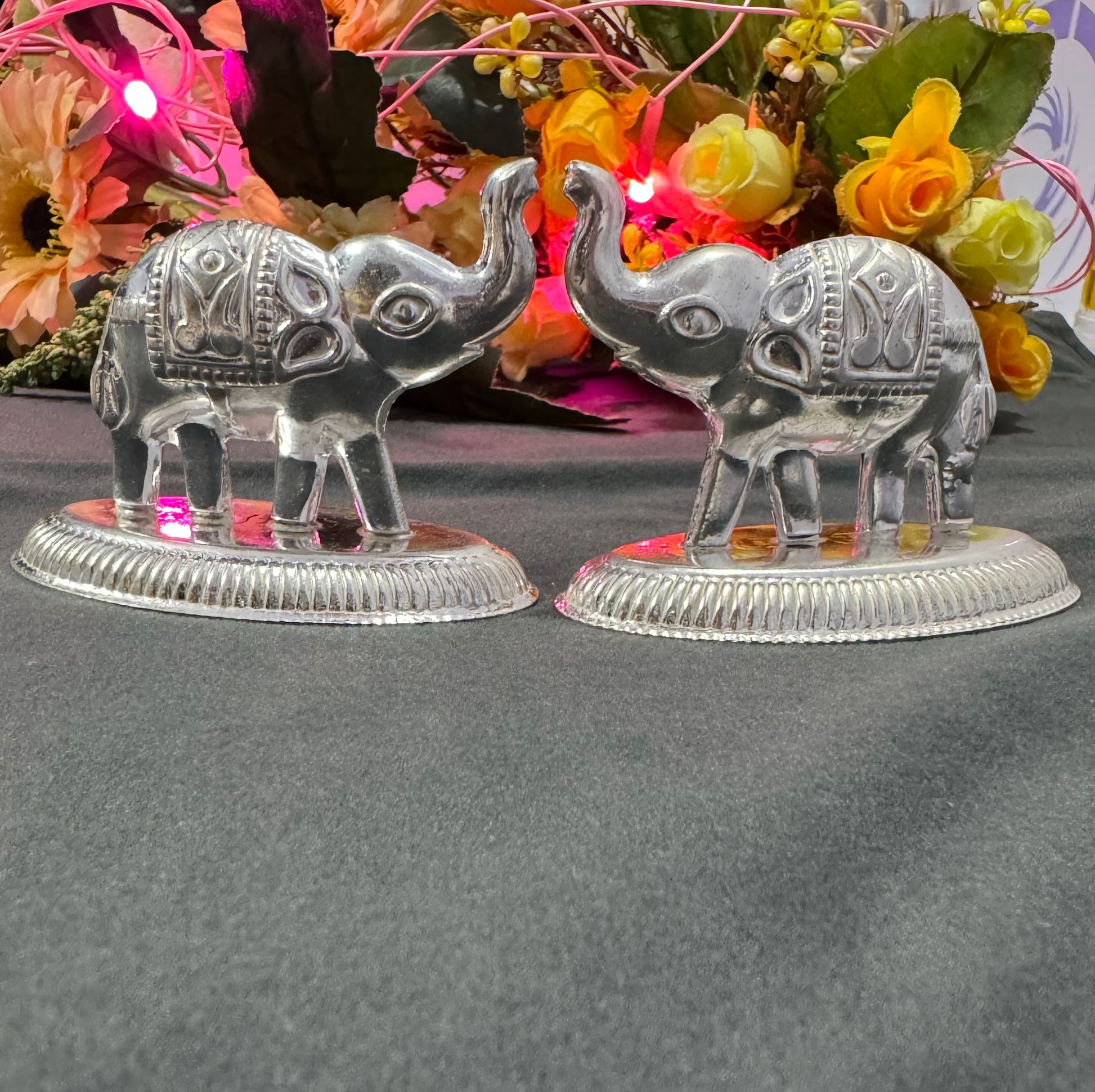 Two silver elephant figurines on a dark surface with colorful flowers in the background.