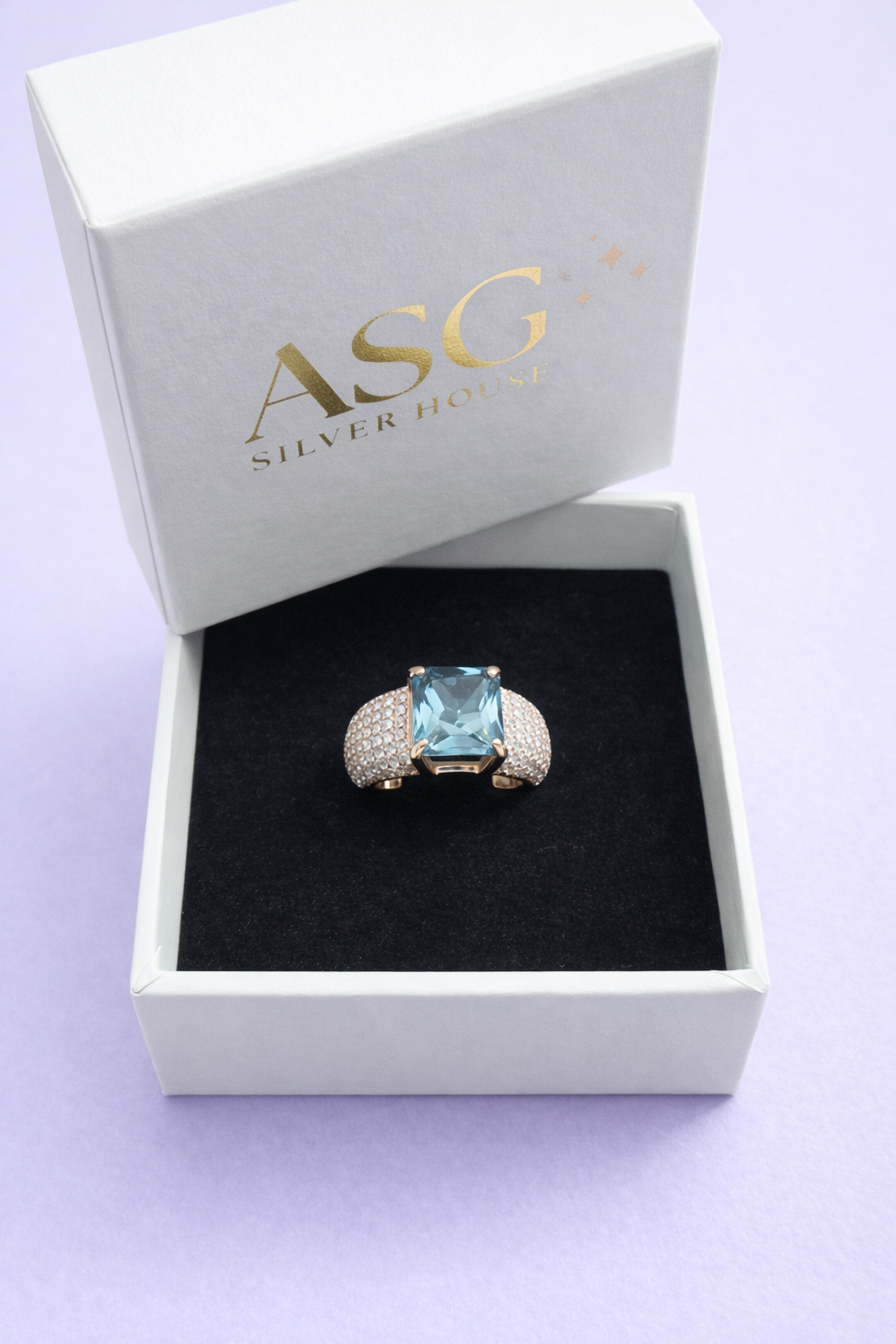 Silver ring with a blue gemstone in an open box labeled 'ASG Silver House' on a light purple background.